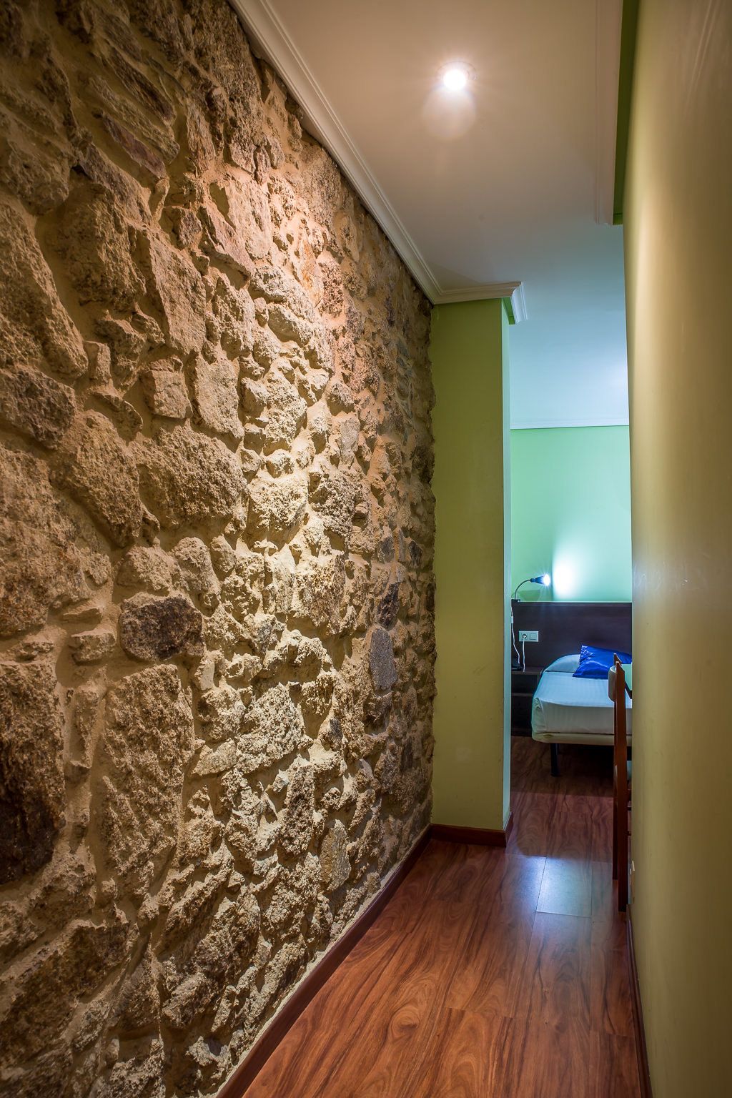 Un couloir étroit, avec un mur en pierre d’un côté et un plancher en bois. On aperçoit un lit avec un oreiller bleu au loin.