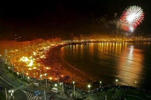 A nighttime view of a coastal city with a large crowd gathered along the waterfront, a bridge, and fireworks in the sky. - Hostal Hotil (A Coruña)