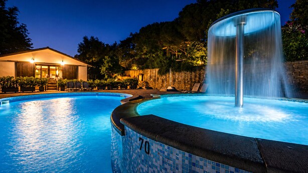 A swimming pool with a water feature, a building with a roof and windows, and a stone wall.