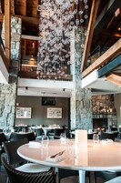 Restaurante com lareira de pedra, candelabro de cristal pendente e várias mesas de jantar dispostas com toalhas brancas e copos de vinho.