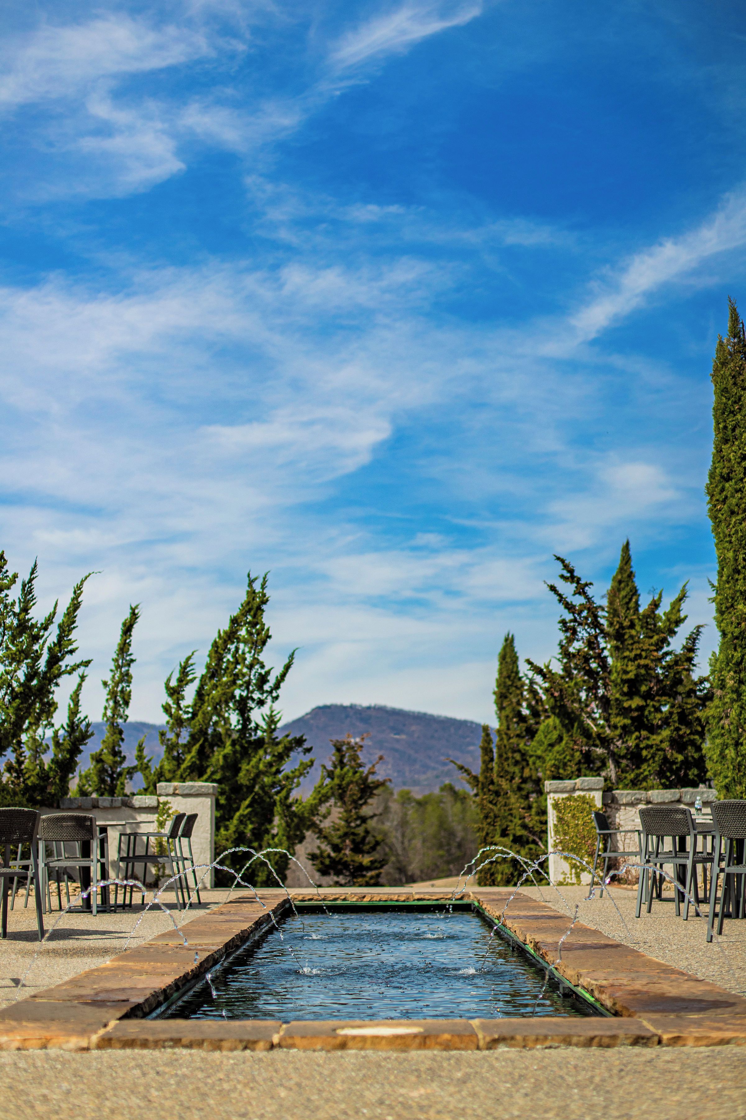 A rectangular pool with a stone border, surrounded by chairs and tables, with mountains in the background.