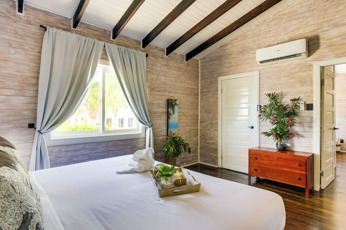 Een slaapkamer met een groot bed, een houten dressoir, een potplant en een raam met gordijnen.