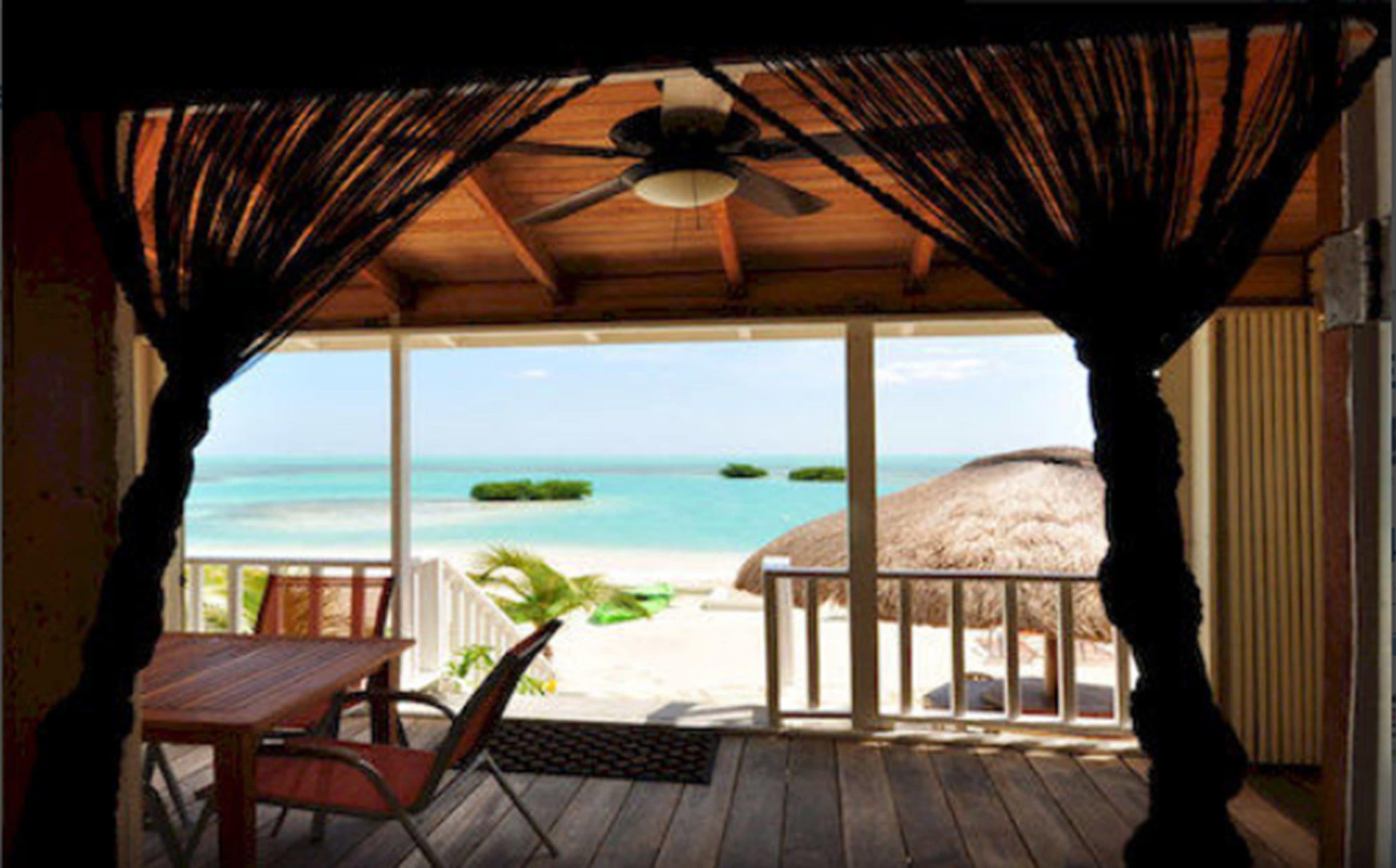A wooden deck with a table and chairs, overlooking a beach with a thatched-roof structure.