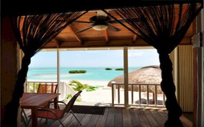 A wooden deck with a table and chairs, overlooking a beach with a thatched-roof structure. - Shaka Caye - All inclusive (Little Frenchman Caye)