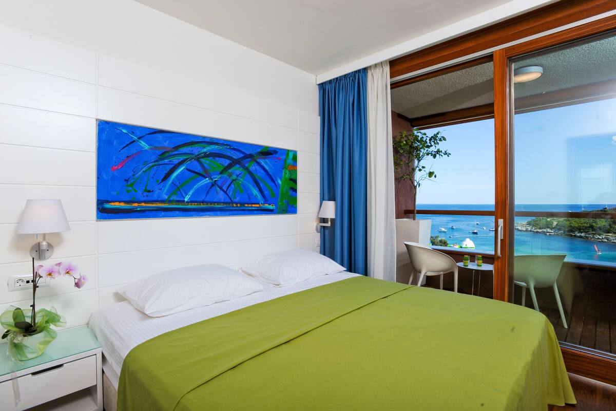 A hotel room with a bed, a painting, a window with a view of the sea, and a plant on the windowsill.