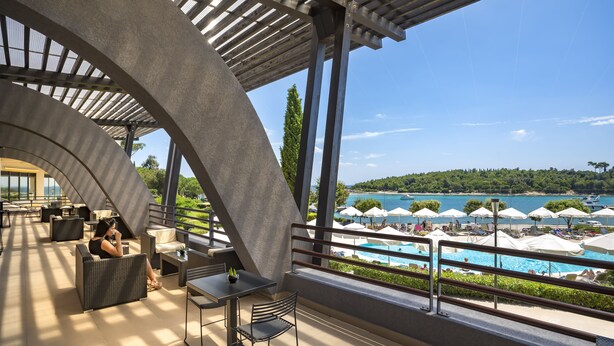 A modern outdoor lounge area with a pool view, featuring a curved ceiling structure, outdoor seating, and a railing overlooking the pool. - Maistra Select All Suite Island Hotel Istra  (Rovinj)