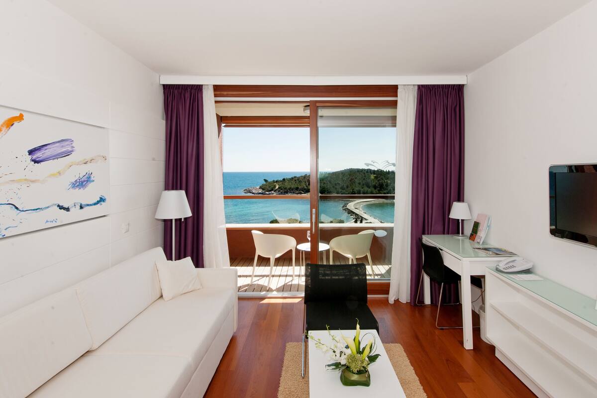 A modern room with a white sofa, a desk with a chair, a TV, and a view of the sea through large windows.