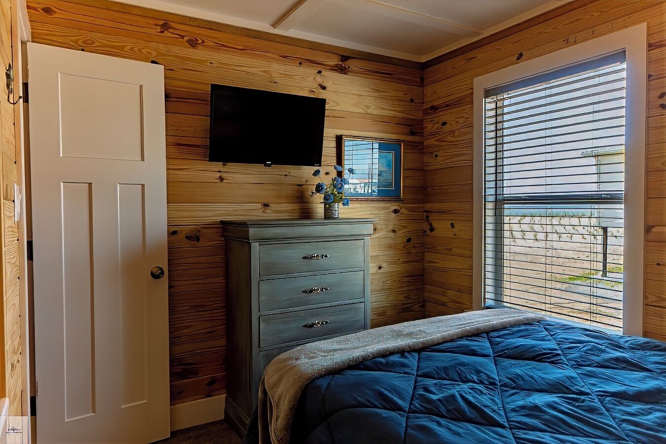 A bedroom with wooden walls, a bed with a blue blanket, a dresser, a TV mounted on the wall, and a window with blinds.