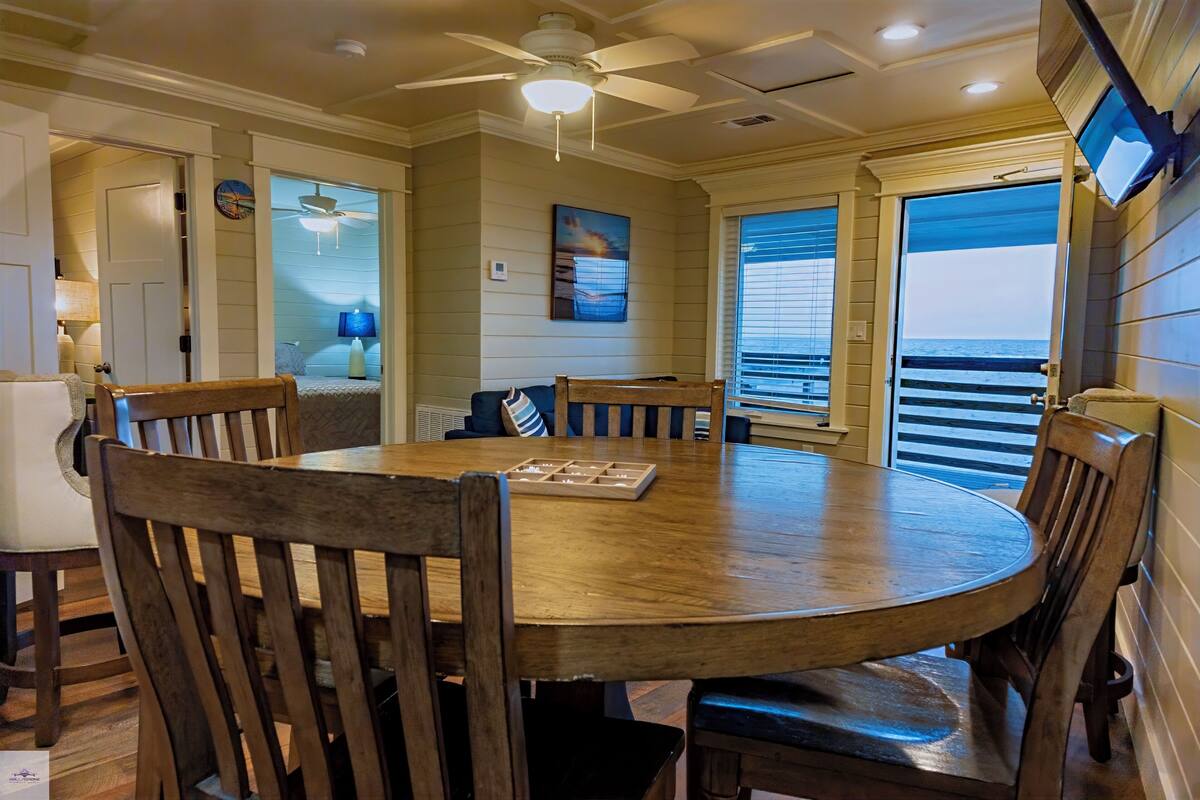 A dining area with a round wooden table, multiple chairs, a ceiling fan, and a view of the ocean through large windows.