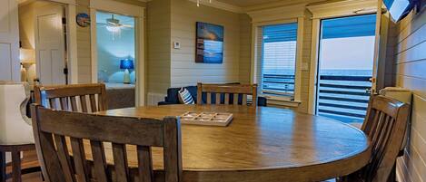 A dining area with a round wooden table, multiple chairs, a ceiling fan, and a view of the ocean through large windows.