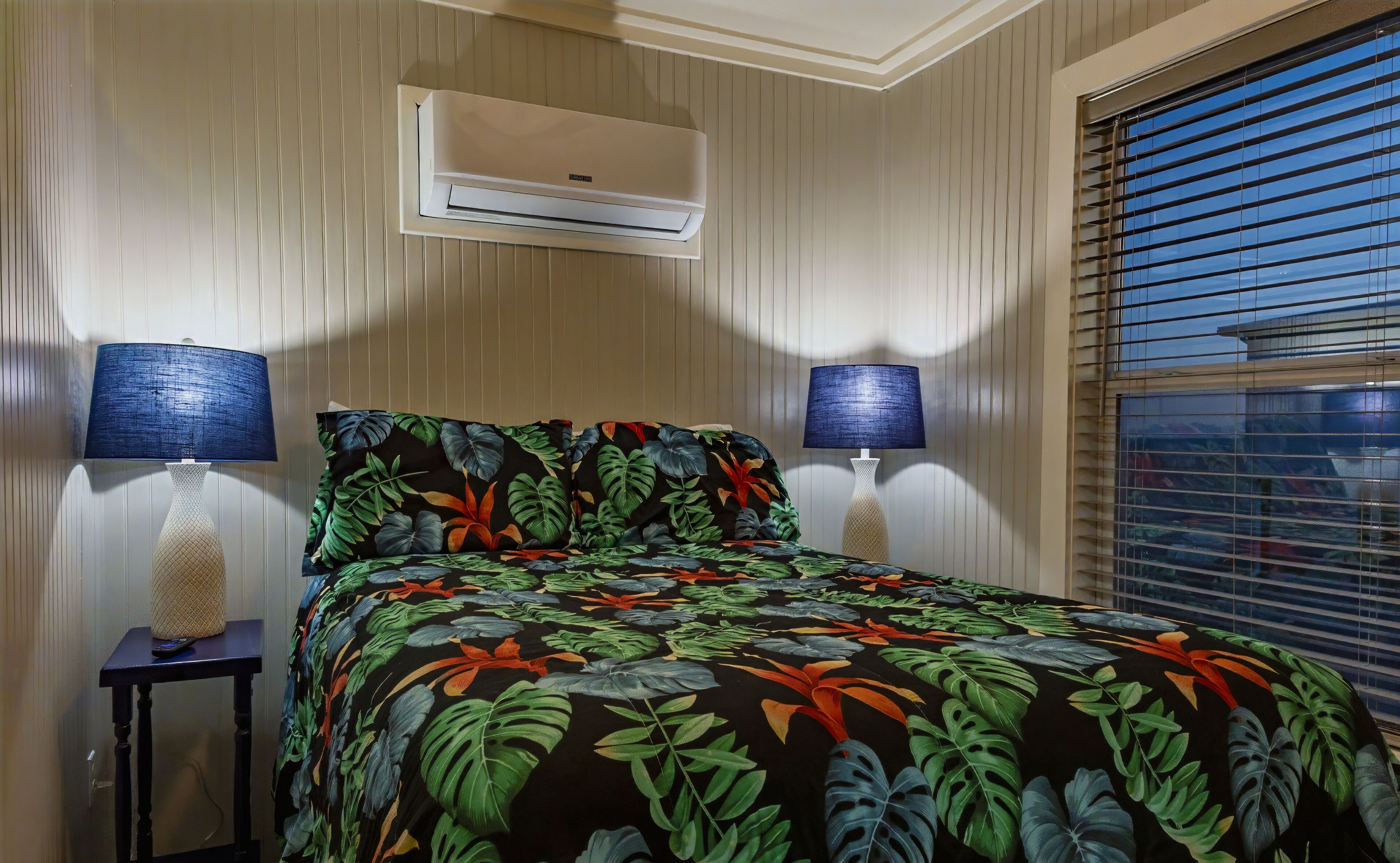 Un dormitorio con cama, dos lámparas de noche, aire acondicionado y una ventana con persianas.
