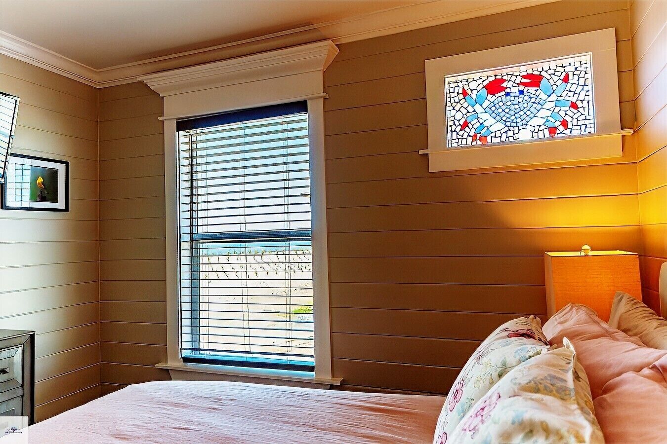 Une chambre avec une grande fenêtre, un lit recouvert d’une literie à motifs floraux, une lampe de chevet et un vitrail au-dessus du lit.
