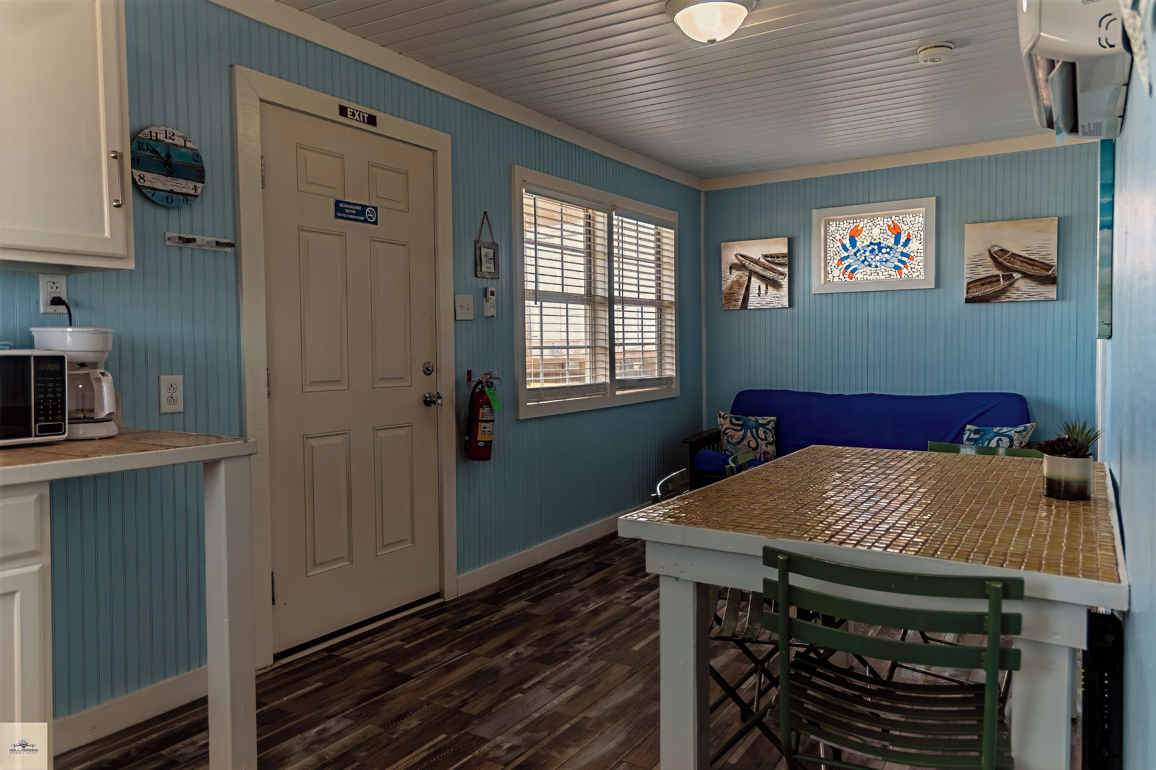Una habitación con piso de madera, una puerta blanca con el letrero 'EXIT', una ventana con persianas, un sofá azul, una mesa con sillas y un reloj de pared.