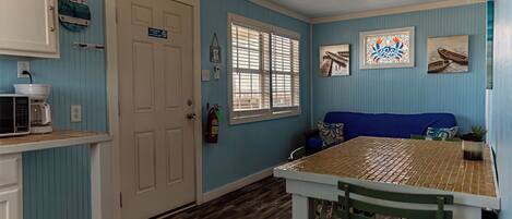 Una habitación con piso de madera, una puerta blanca con el letrero 'EXIT', una ventana con persianas, un sofá azul, una mesa con sillas y un reloj de pared.