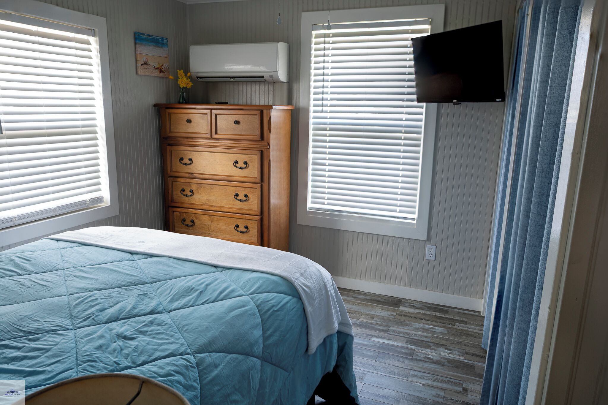 A bedroom with a bed, a wooden dresser, a television mounted on the wall, and a window with blinds.