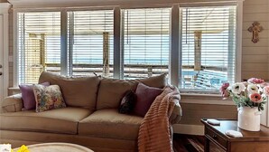 A cozy living room with a beige sofa, a wooden ceiling, and a view of the beach through large windows. - Blue Dolphin Inn and Cottages (Grand Isle)