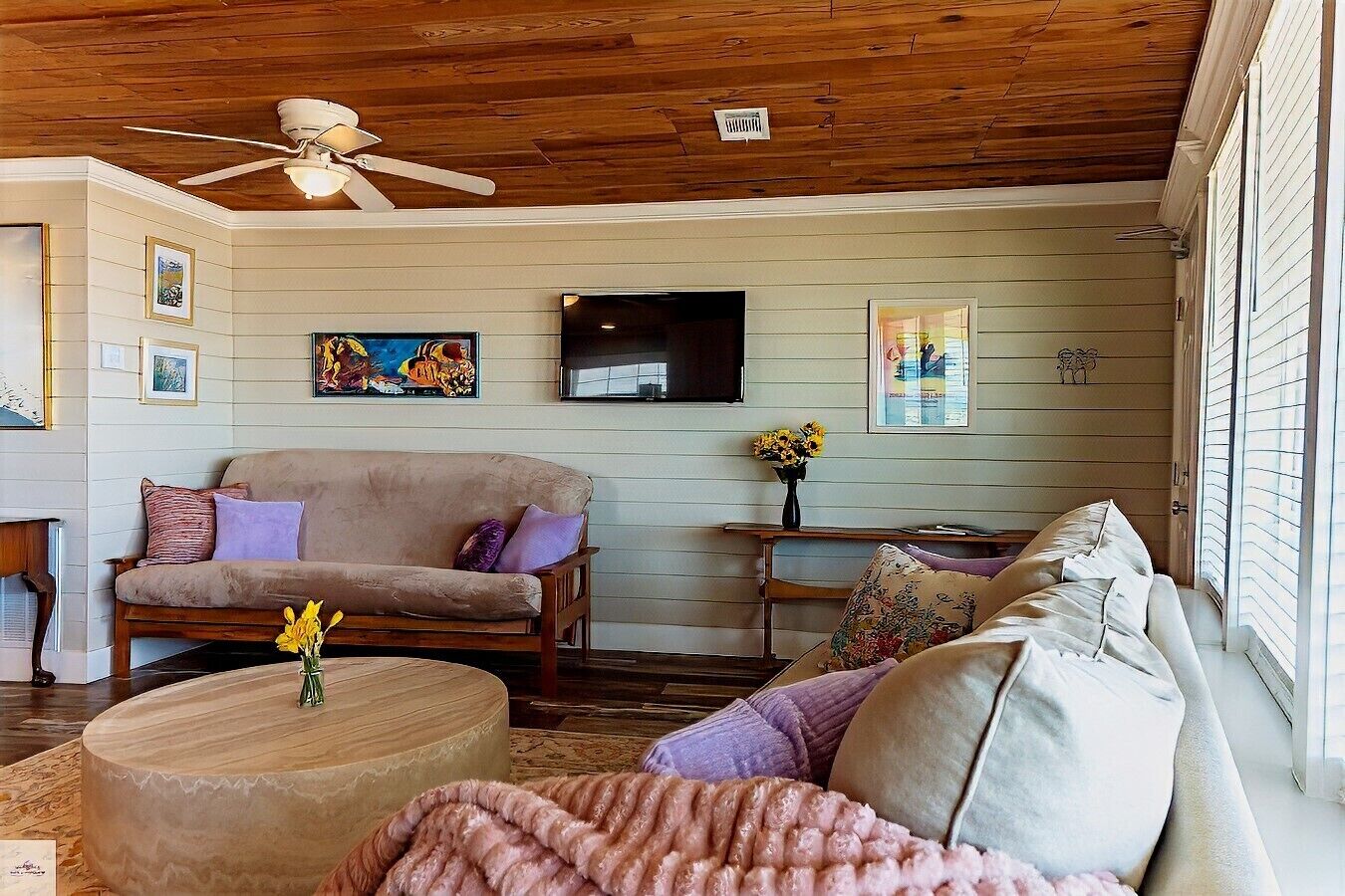 A cozy living room with a wooden ceiling, a beige sofa, a round coffee table, and a television mounted on the wall.
