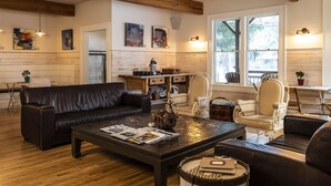 Una sala de estar acogedora con un sofá de cuero oscuro, una mesa de centro de madera y un sillón color beige. Se observan vigas de madera en el techo, una chimenea y un ventanal con vistas al exterior.