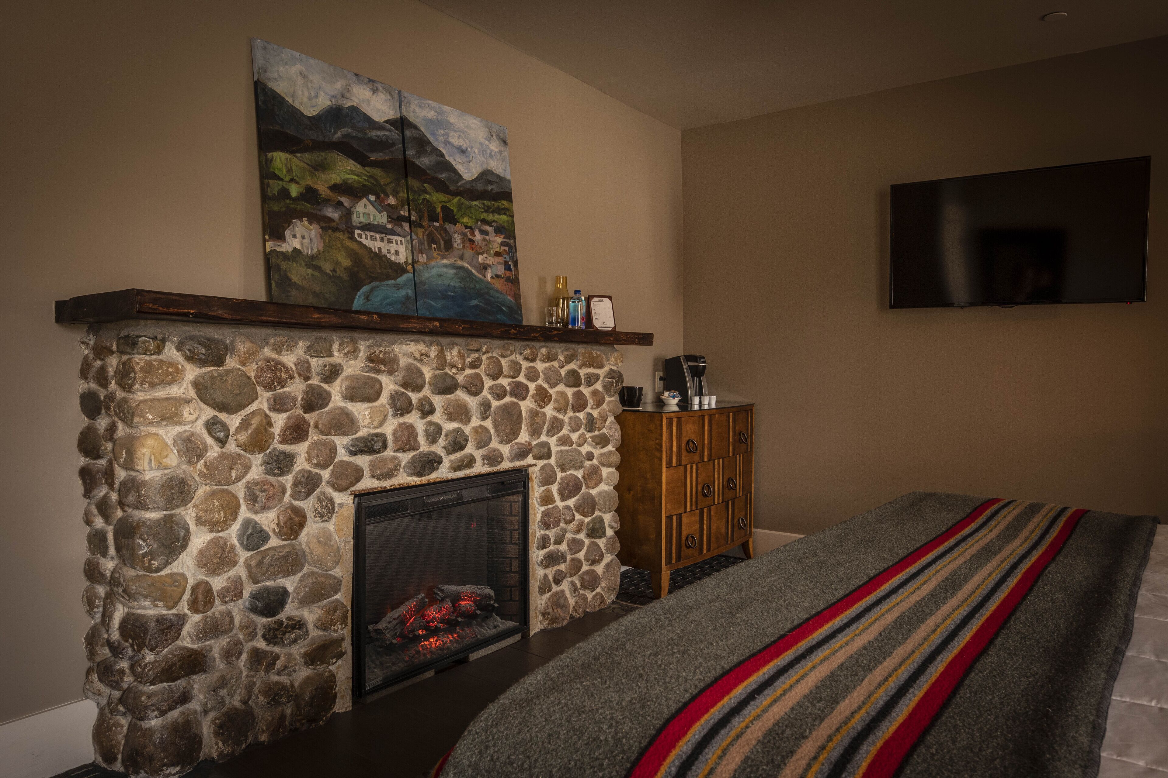 A stone fireplace with a painting above it, a television mounted on the wall, and a wooden cabinet to the right.