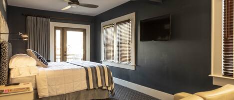 A bedroom with a bed, a desk, a chair, a ceiling fan, and a TV mounted on the wall.