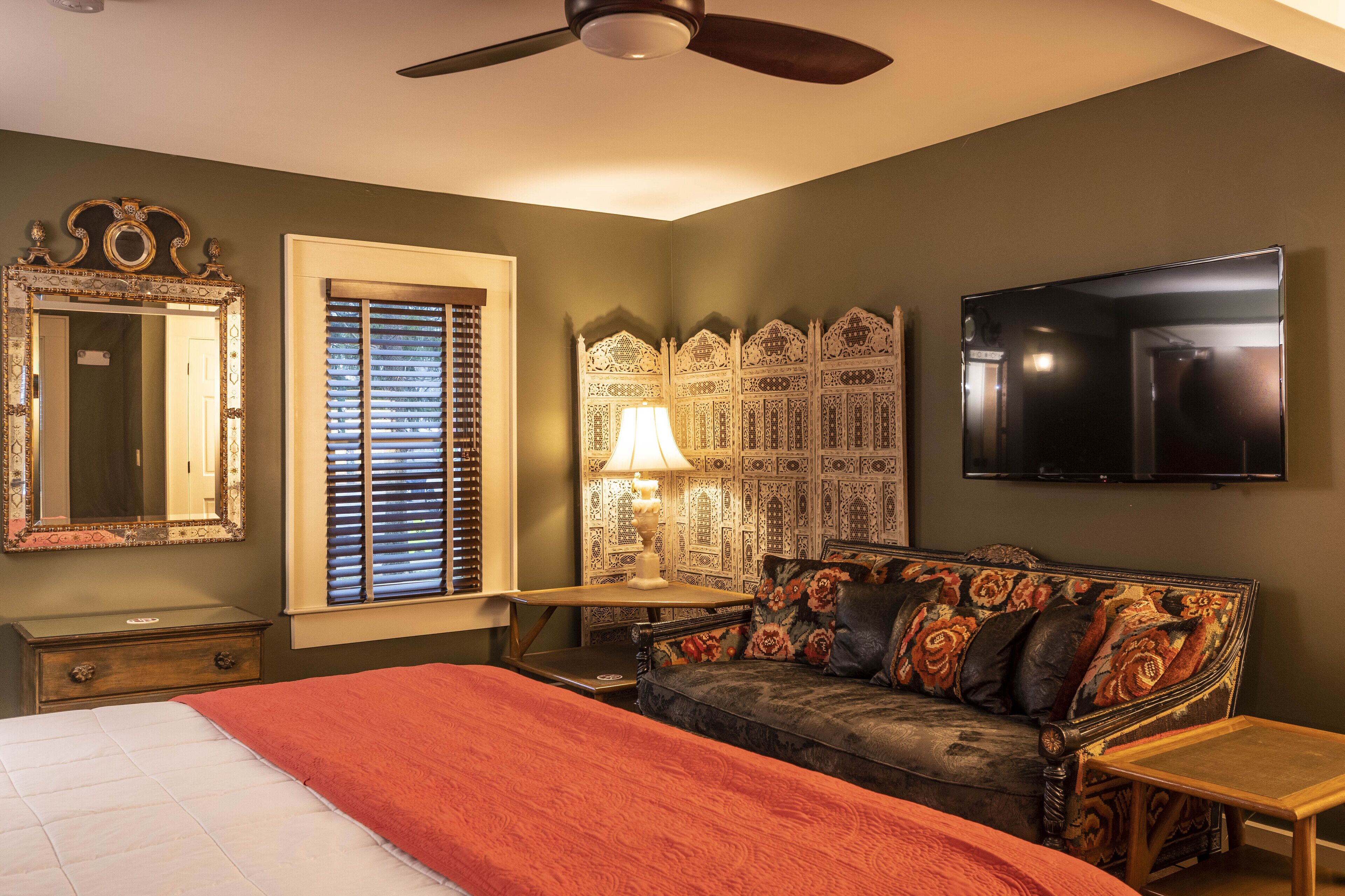 A bedroom with a bed, a nightstand, a dresser, a sofa, a TV, a ceiling fan, and a decorative wall panel.