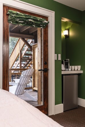 Una habitación con puerta de madera que da a una terraza exterior, una silla blanca y una zona de cocina con cafetera y tazas.