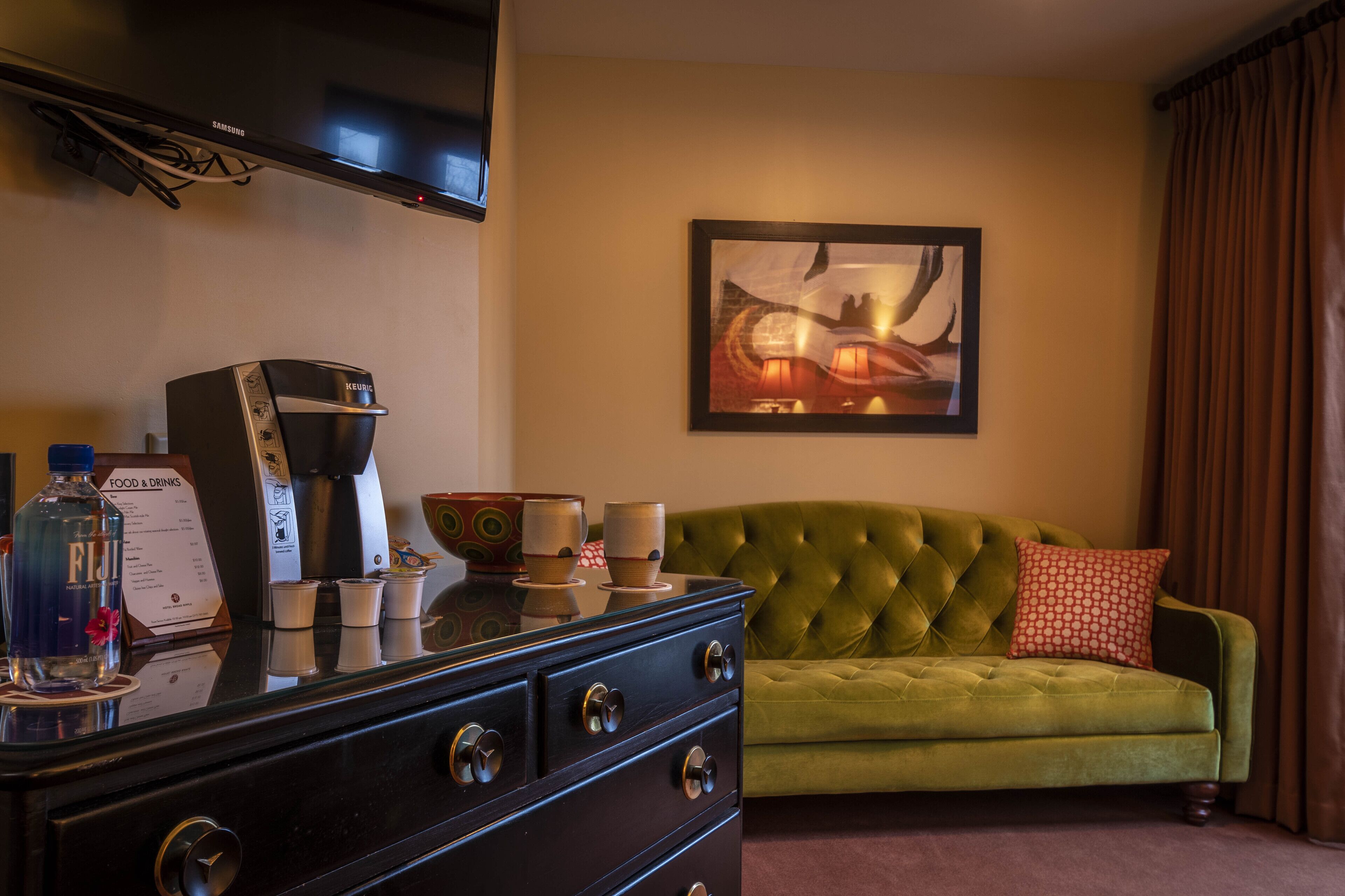 Quarto de hotel com sofá verde, máquina de café, pintura abstrata emoldurada e TV Samsung na parede.