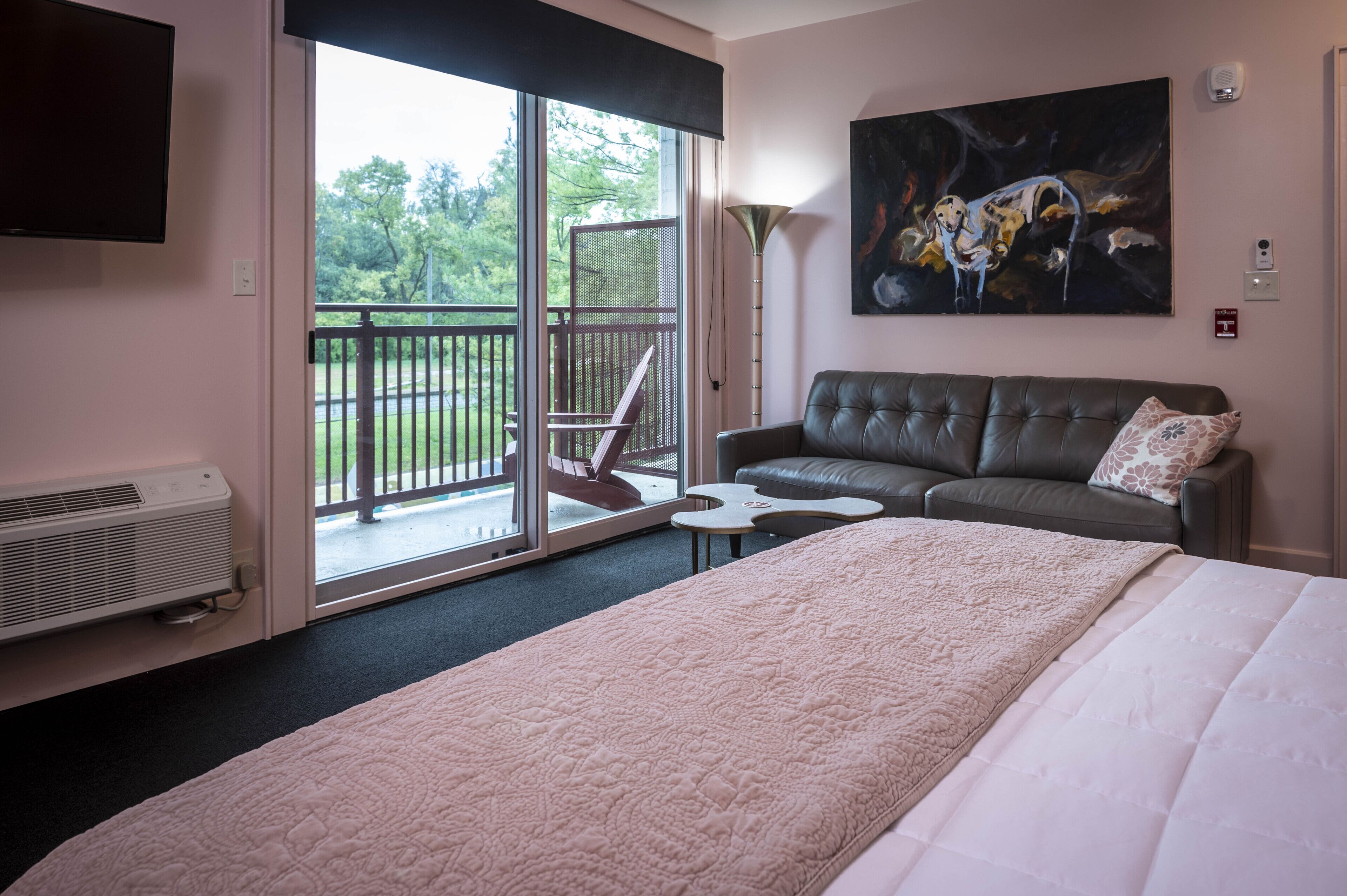 A hotel room with a large bed, a sofa, a small table, a TV, and a balcony with a chair.