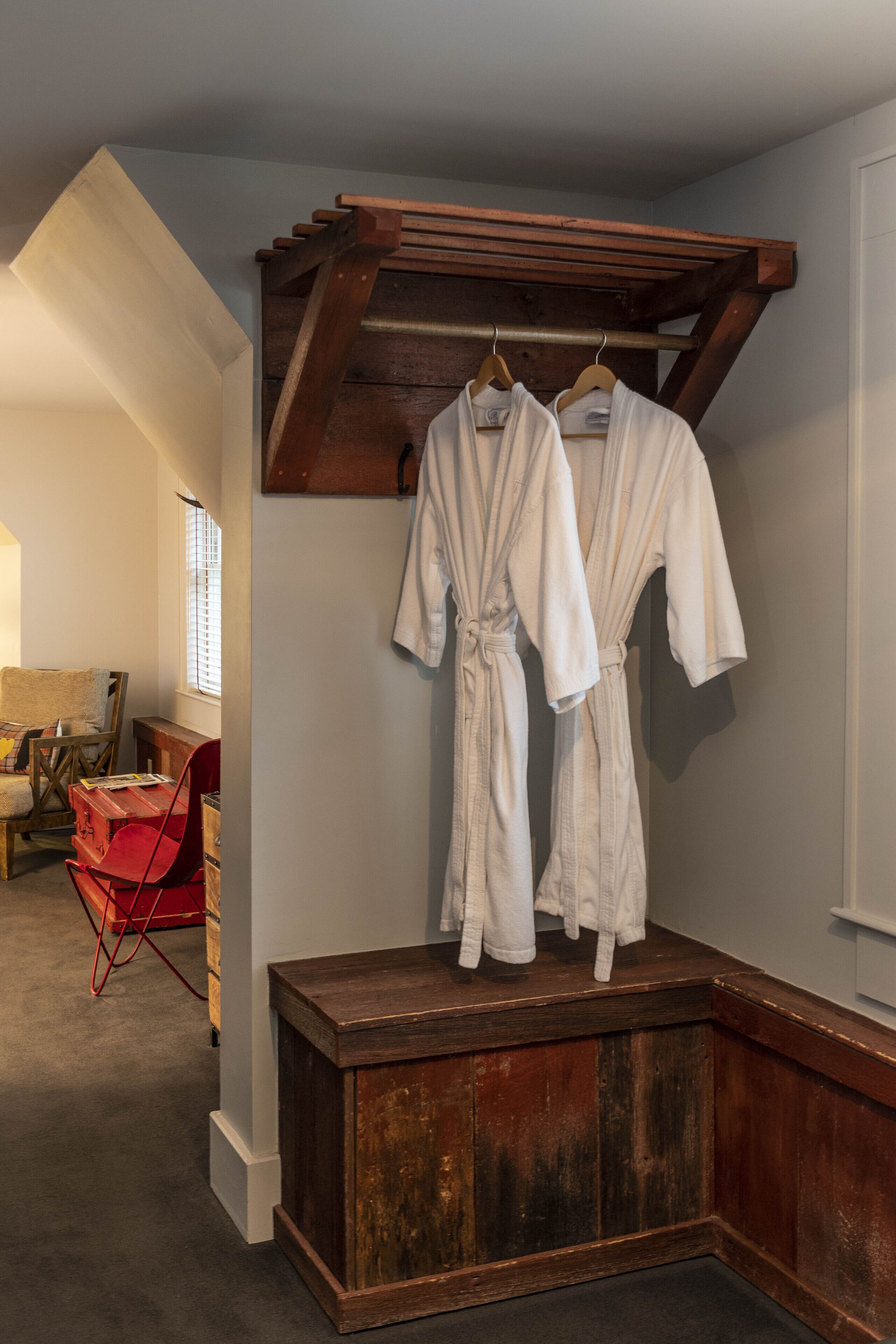 A room with a wooden bench, two white robes hanging on a wooden rack, a red chair, and a small table with a lamp.