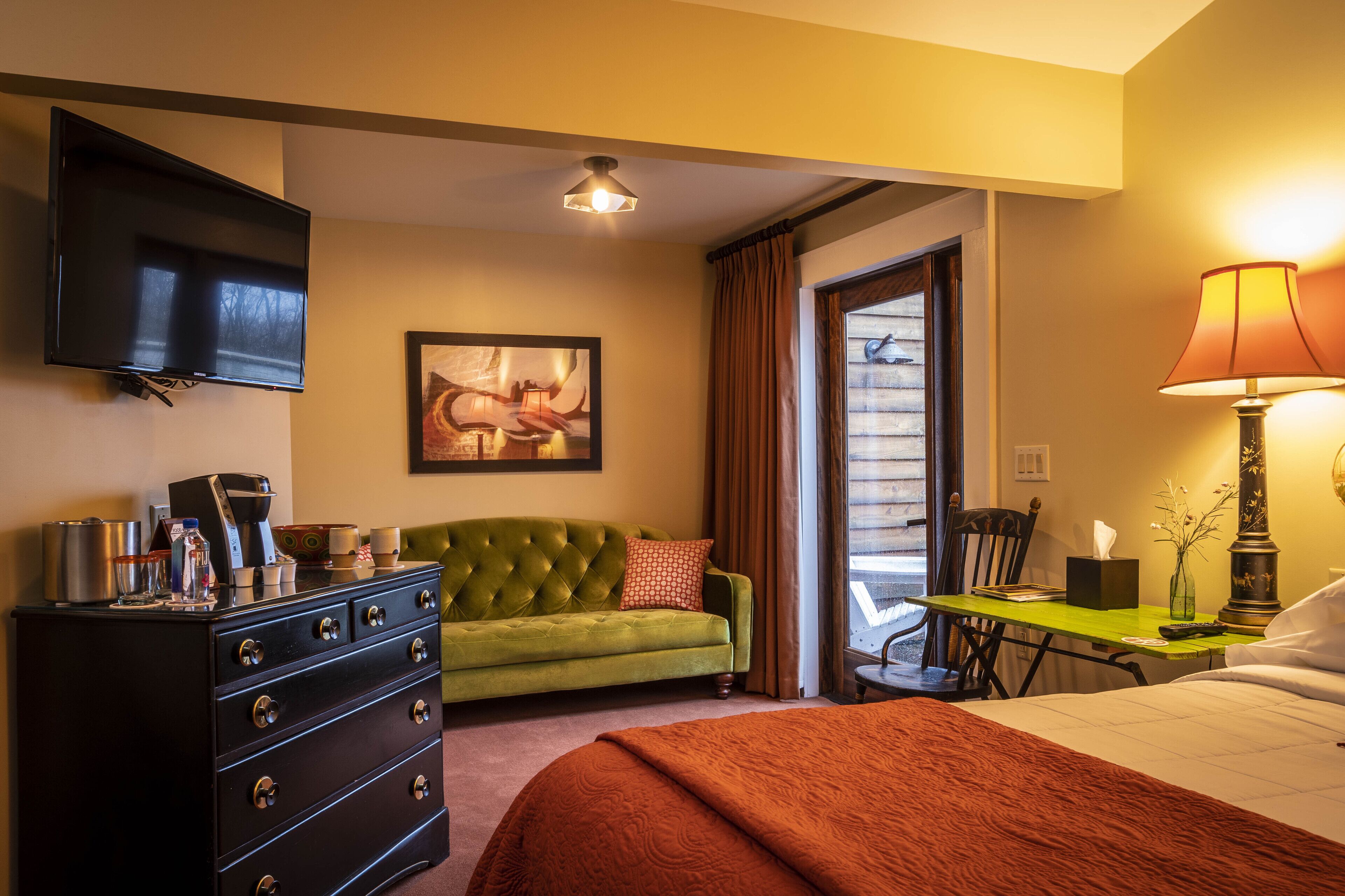 Una habitación de hotel con sofá verde, un televisor de pantalla plana, un aparador, una cama, una mesa con lámpara y una ventana con cortinas.