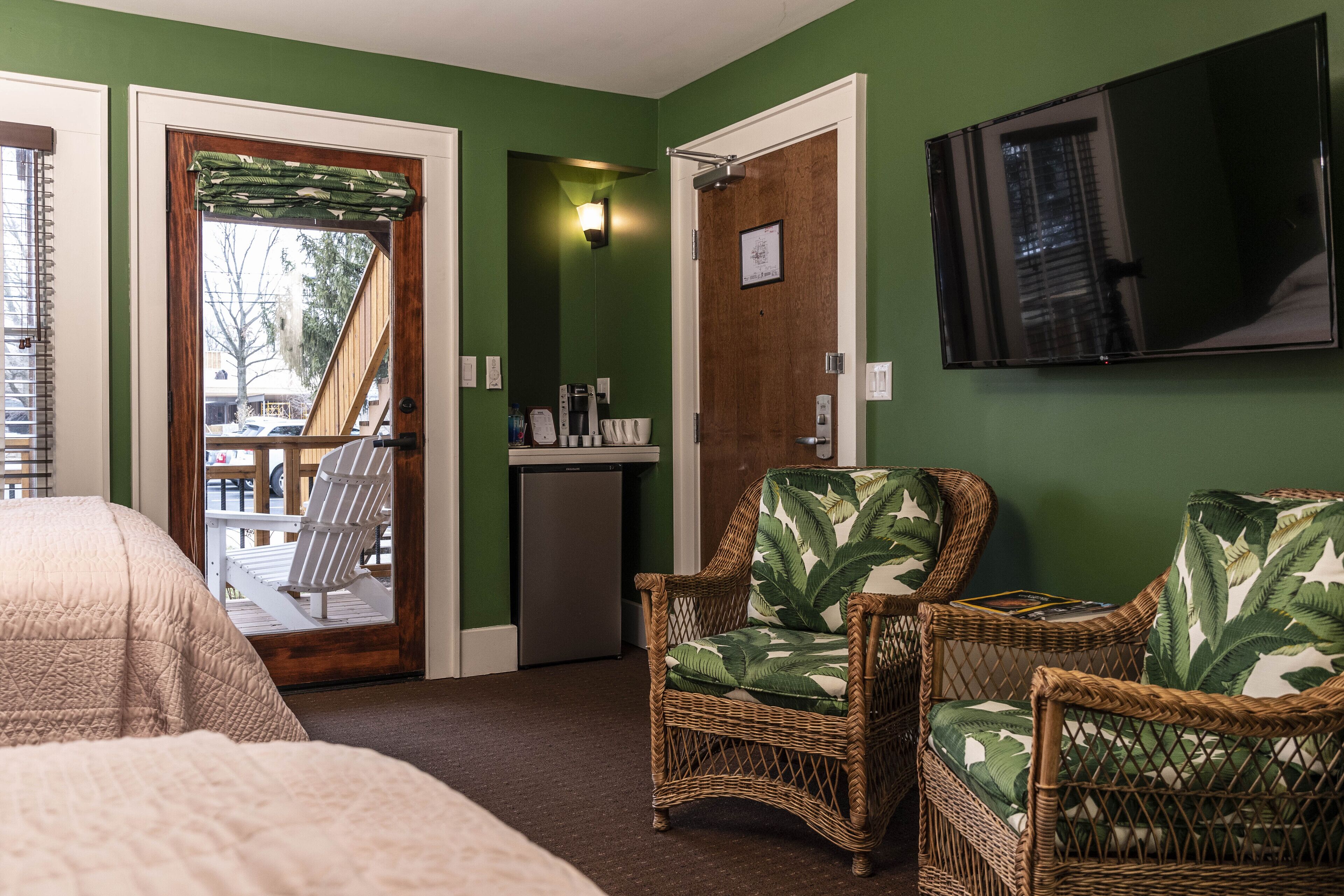 Una camera d'hotel con un letto, due poltrone in vimini con rivestimenti tropicali, un angolo cottura, una televisione e una porta che dà su un'area esterna.