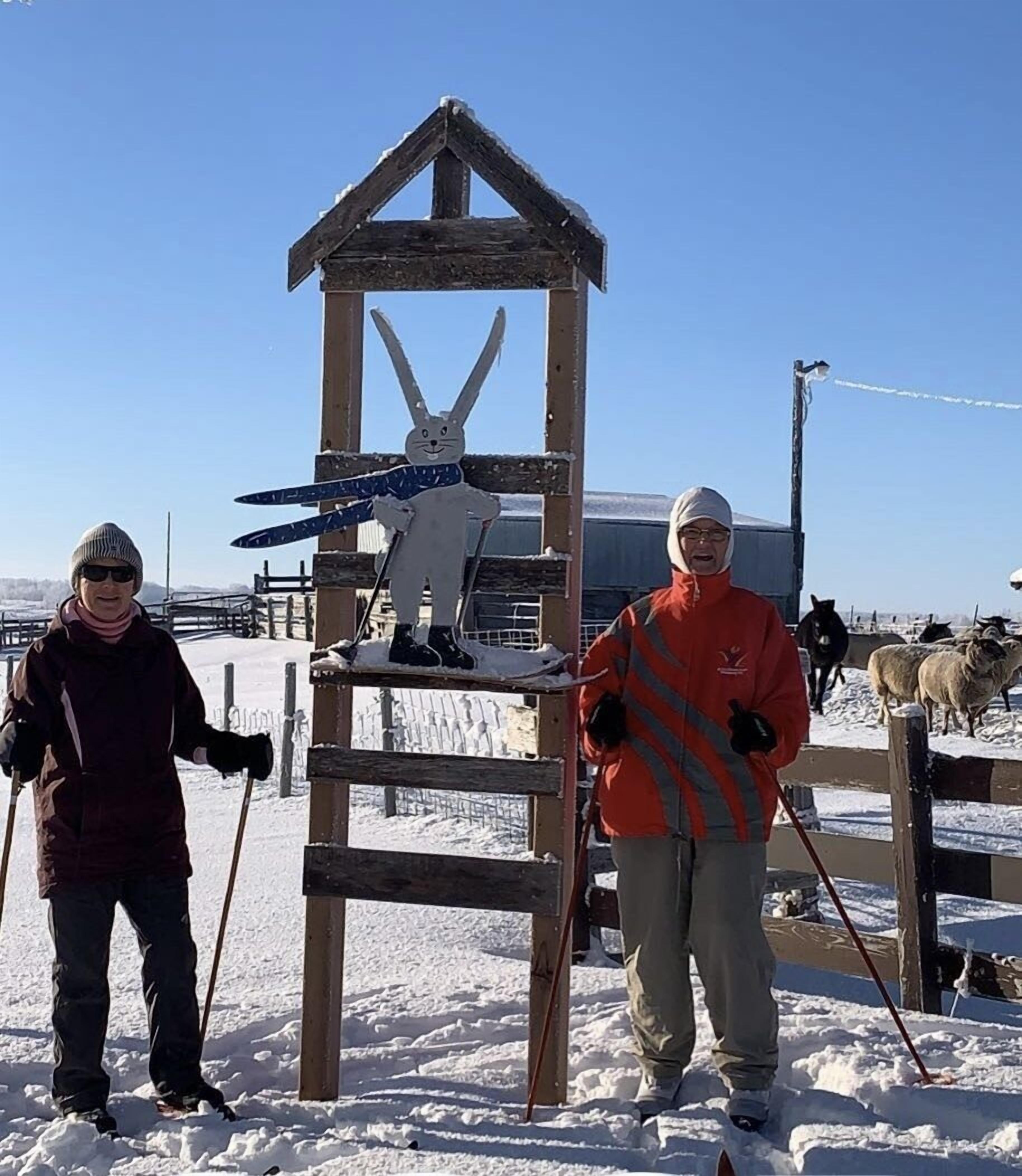 A wooden structure with a snowman figure, a person in a red jacket, and another in a maroon jacket standing in a snowy outdoor area with sheep in the background.