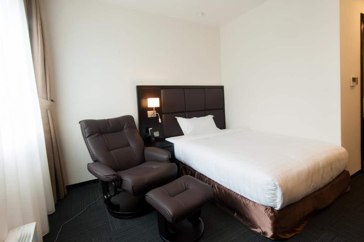 Ein Hotelzimmer mit einem Bett, einem Liegesessel, einem Hocker, einem Nachttisch mit Lampe und einem Fenster mit Vorhängen.