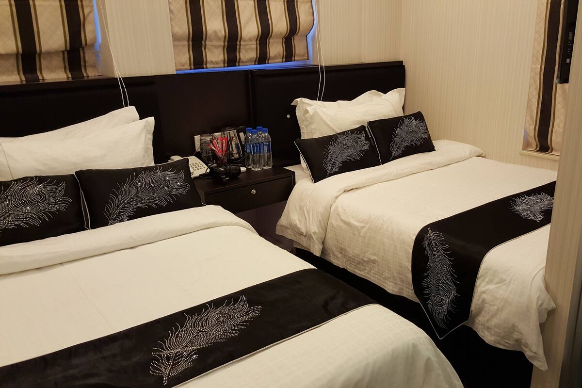Ein Hotelzimmer mit zwei Betten, jeweils mit schwarz-weißer Bettdecke und passenden Kissen. Ein Nachttisch mit Lampe, Telefon und Kamera sind vorhanden.