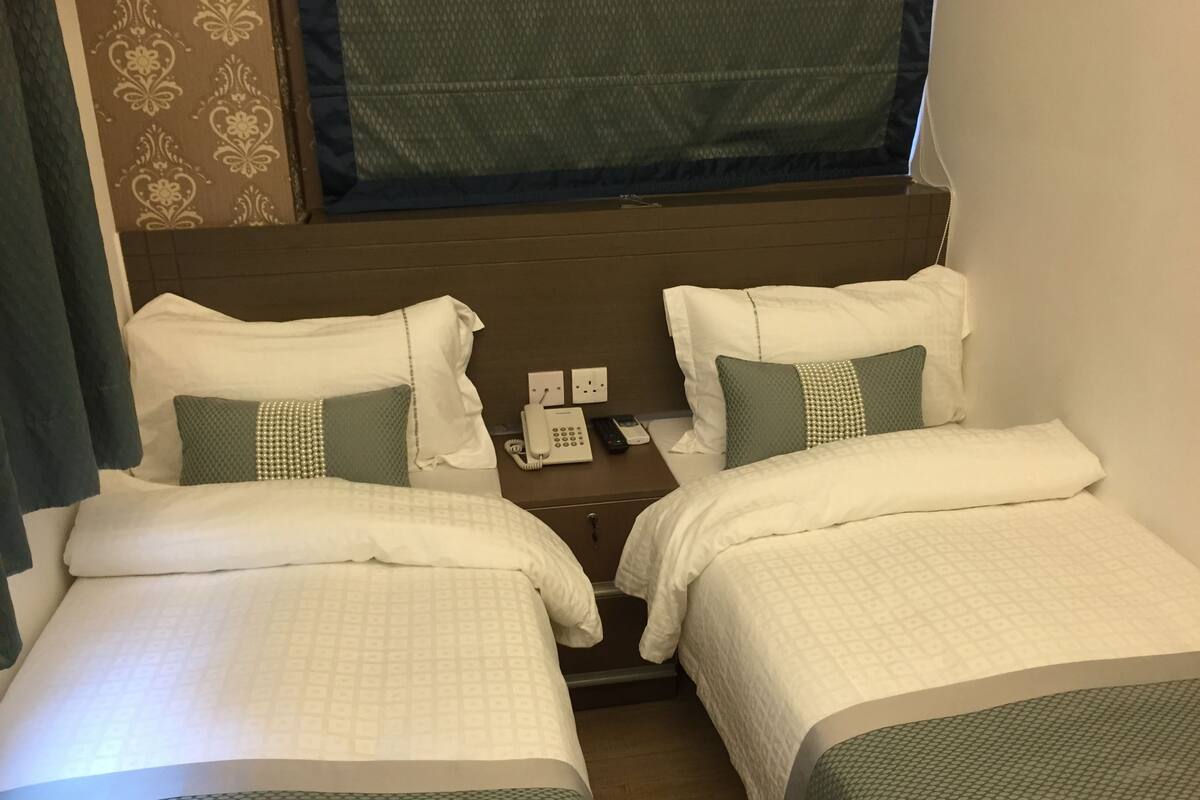 Ein Hotelzimmer mit zwei Betten, jeweils mit weißer Bettwäsche und grünen Akzenten, einem Nachttisch mit Telefon und einem Fenster mit blauer Gardine.