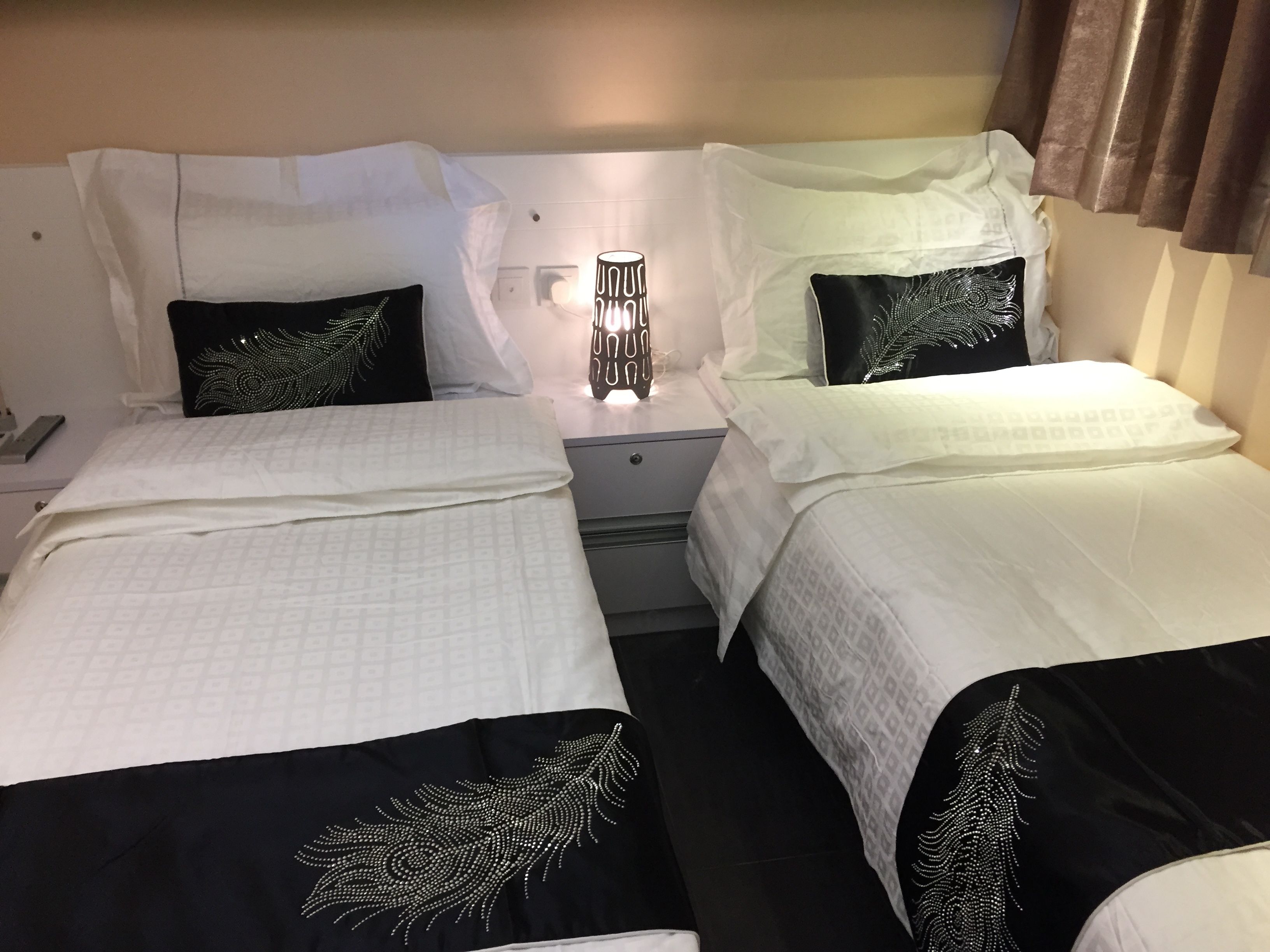 A hotel room with two beds, each with a white bedspread and black pillows with a leaf design. There is a nightstand with a lamp between the beds.