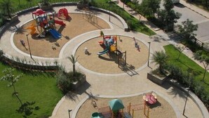Un parc infantil amb diverses zones de jocs, incloent tobogans i estructures per escalar, envoltat d'una zona enjardinada amb camins i vegetació.