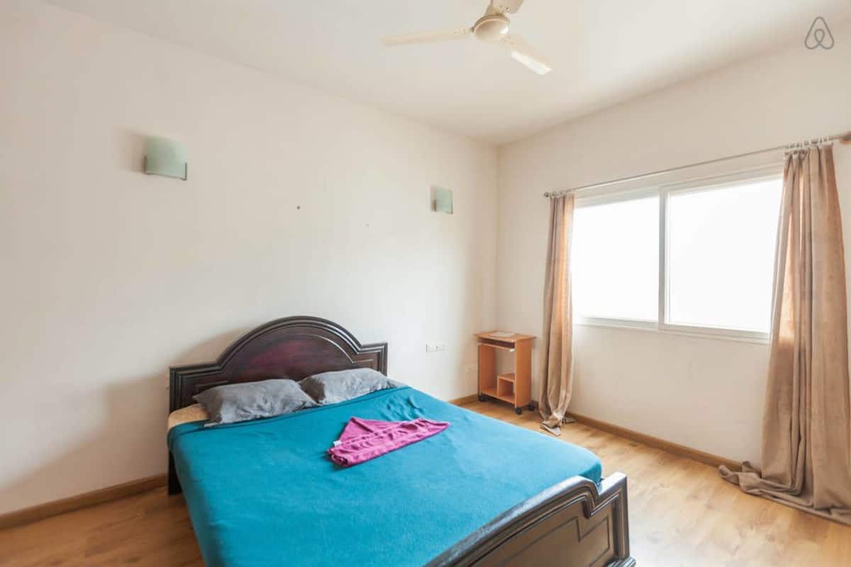 A bedroom with a bed, a ceiling fan, a window with curtains, and a small wooden nightstand.