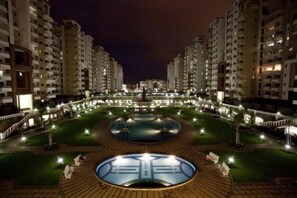 A residential complex with multiple high-rise buildings, a central courtyard with a circular water feature, and a well-lit walkway.