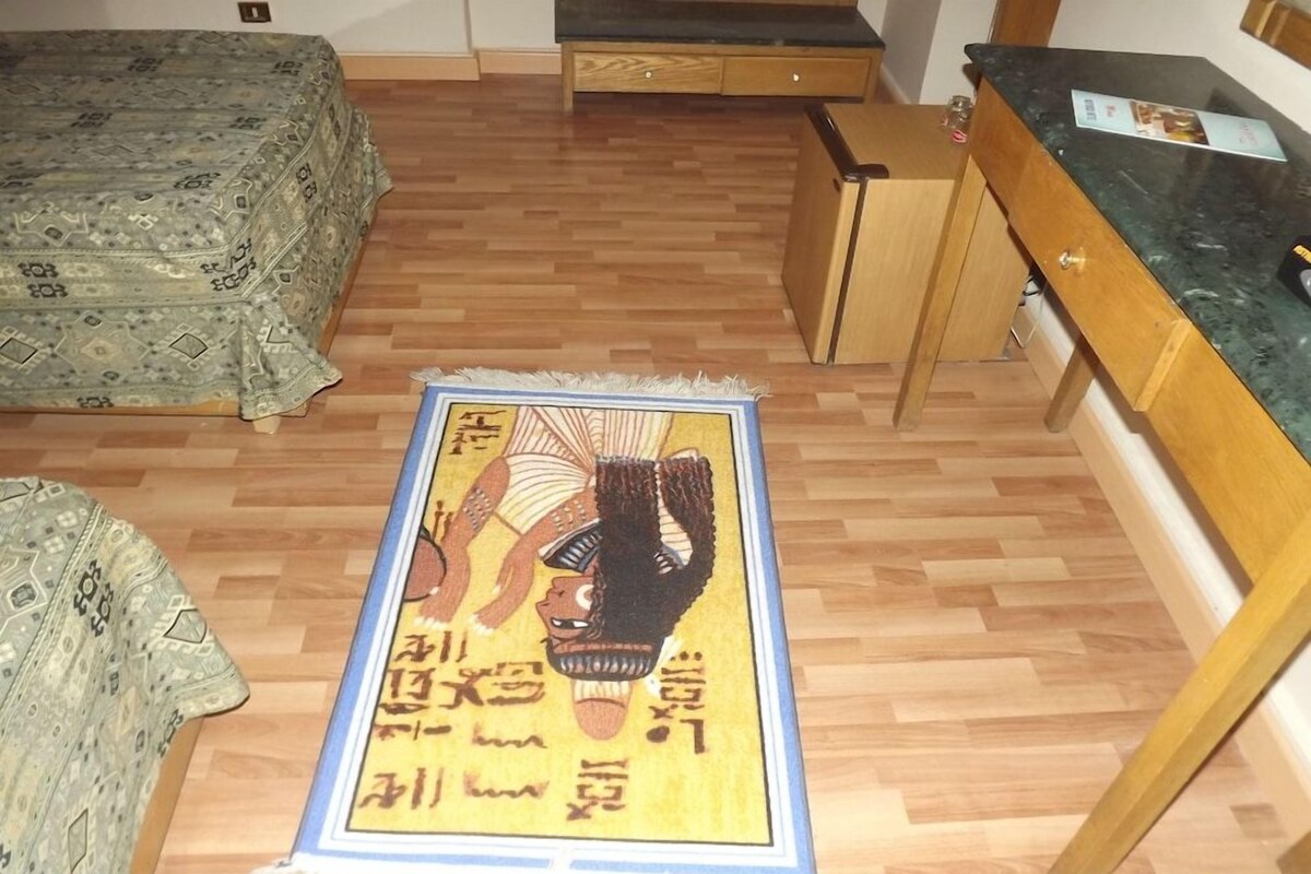 Una habitación con una cama estampada, un pequeño mueble de madera y una zona de cocina con mostrador.