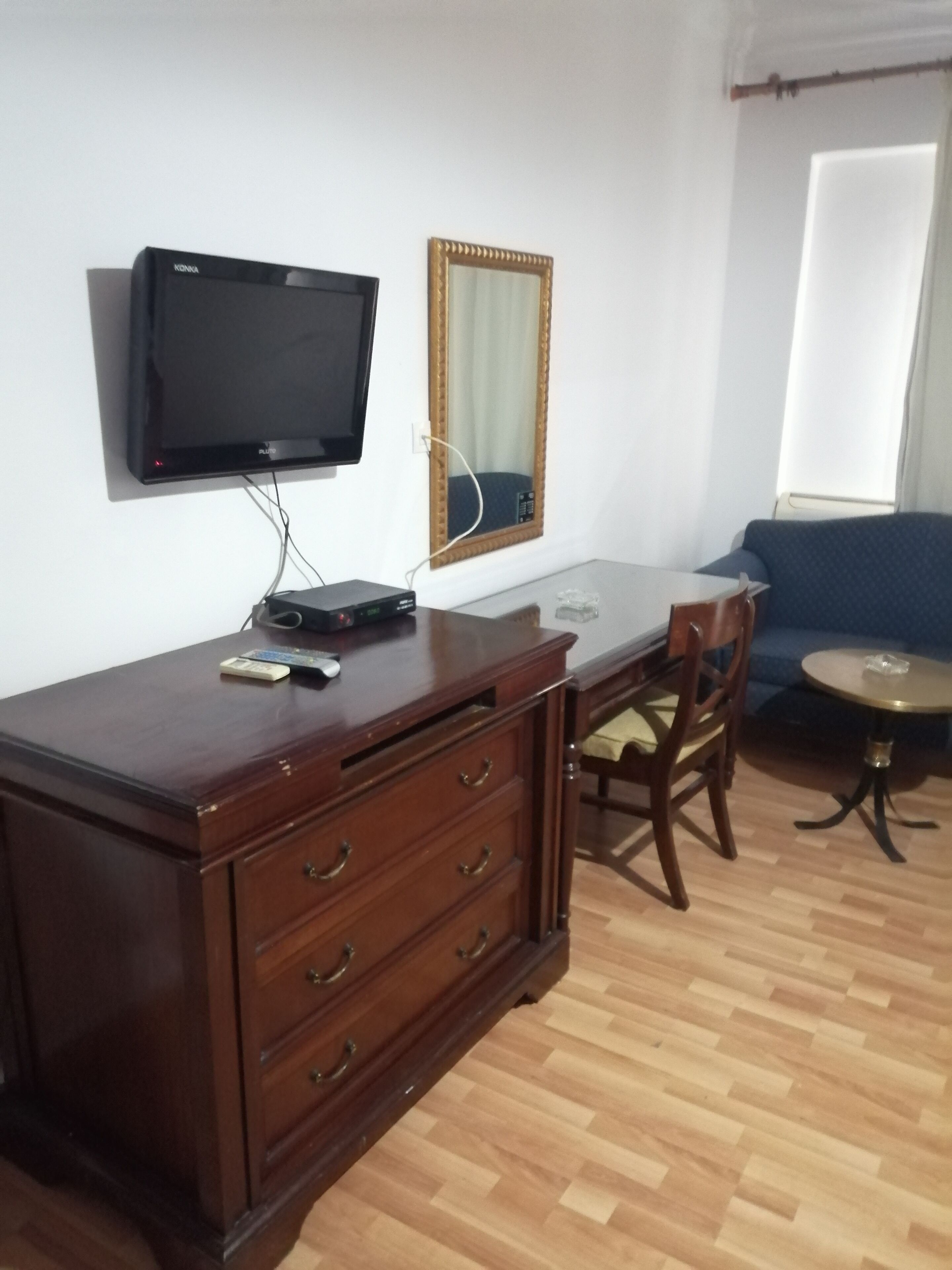 A room with a wooden desk, a television mounted on the wall, a mirror, a chair, and a small round table.
