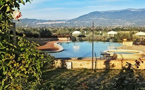 A pool with a curved edge, surrounded by lounge chairs and umbrellas, with a view of distant mountains and a cityscape. - Tenuta del Perugino (Bettona)