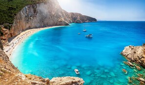 A coastal beach with clear turquoise waters, rocky cliffs, and boats anchored near the shore. - Archontiko (Malevizi)