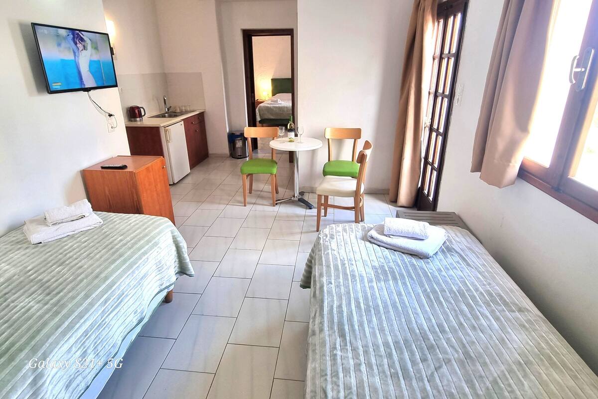 A hotel room with two beds, a desk, a chair, a small table, a TV, and a window with curtains.