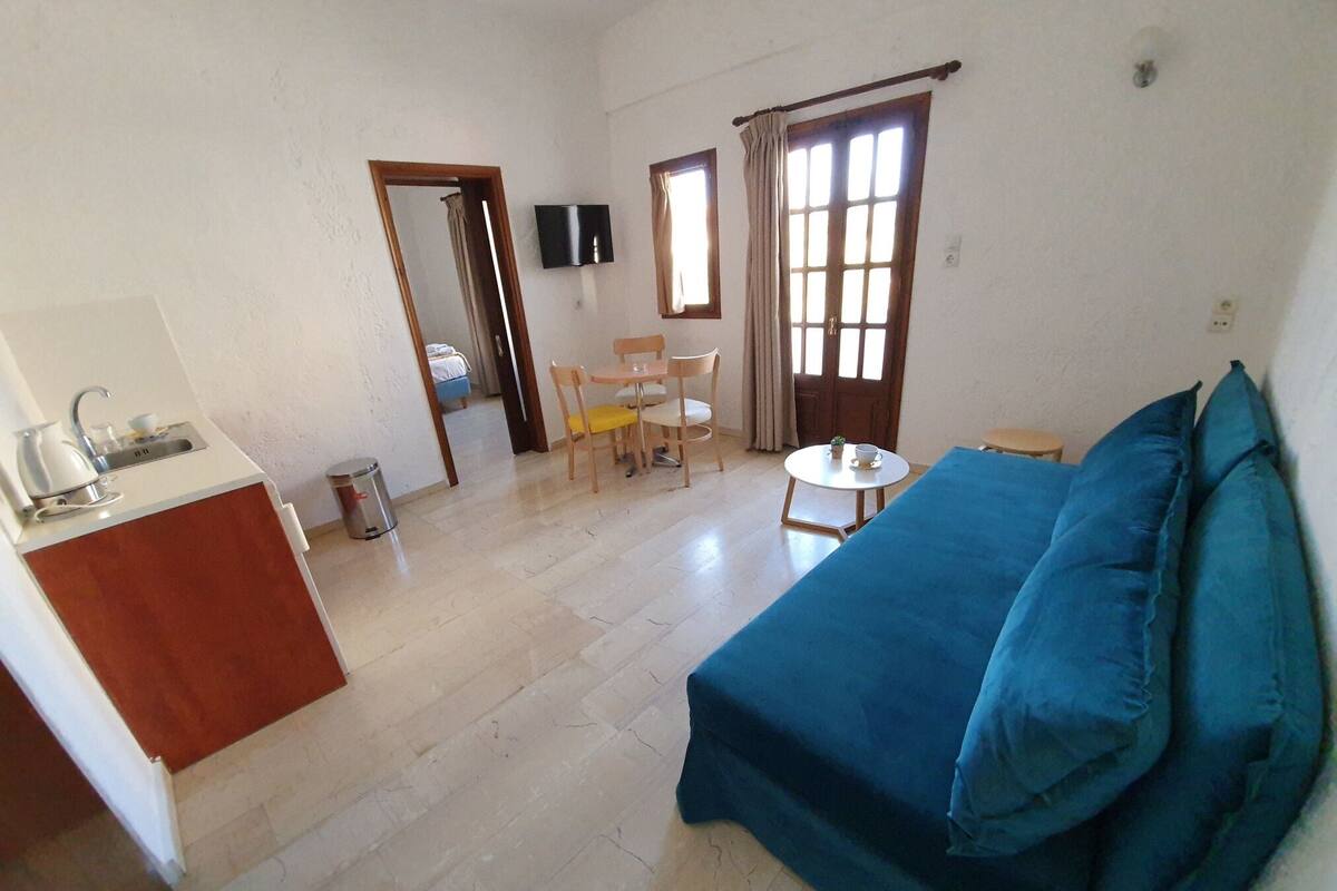 A room with a kitchenette, a sofa, a dining table with chairs, and a small round table with a cup and saucer.