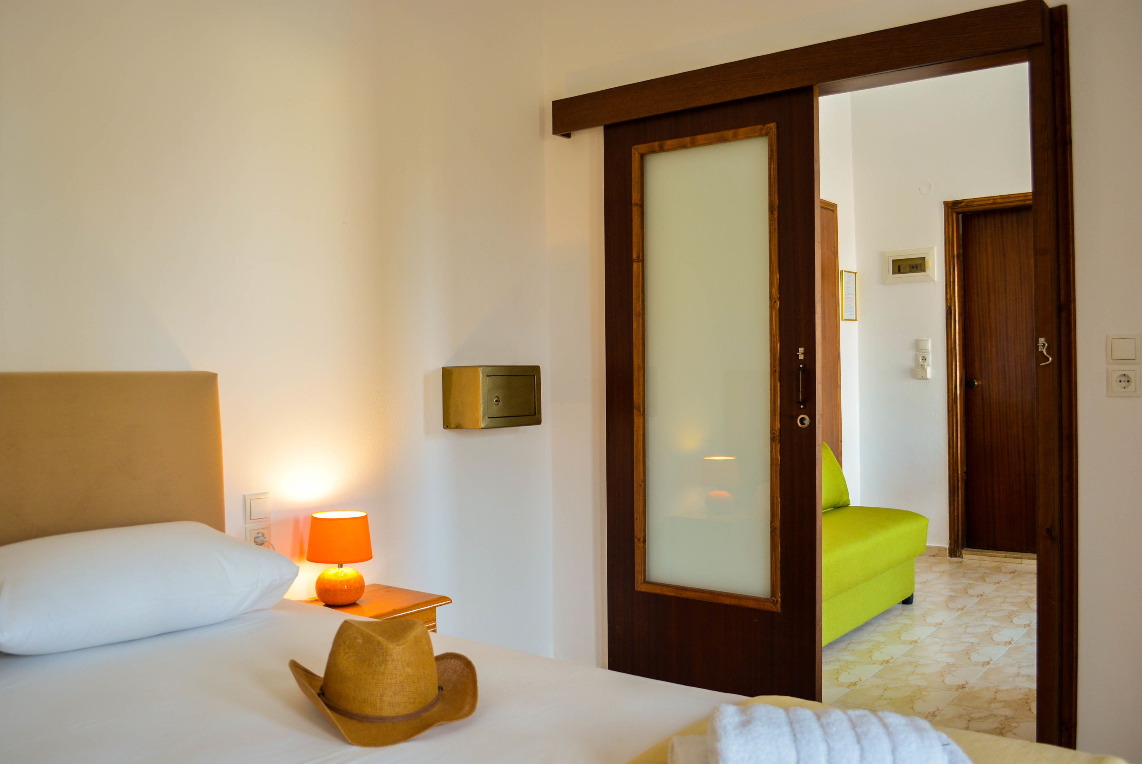 Habitación de hotel con una cama, una mesita de noche con lámpara, una puerta de madera y vista a otra habitación con un sofá verde.