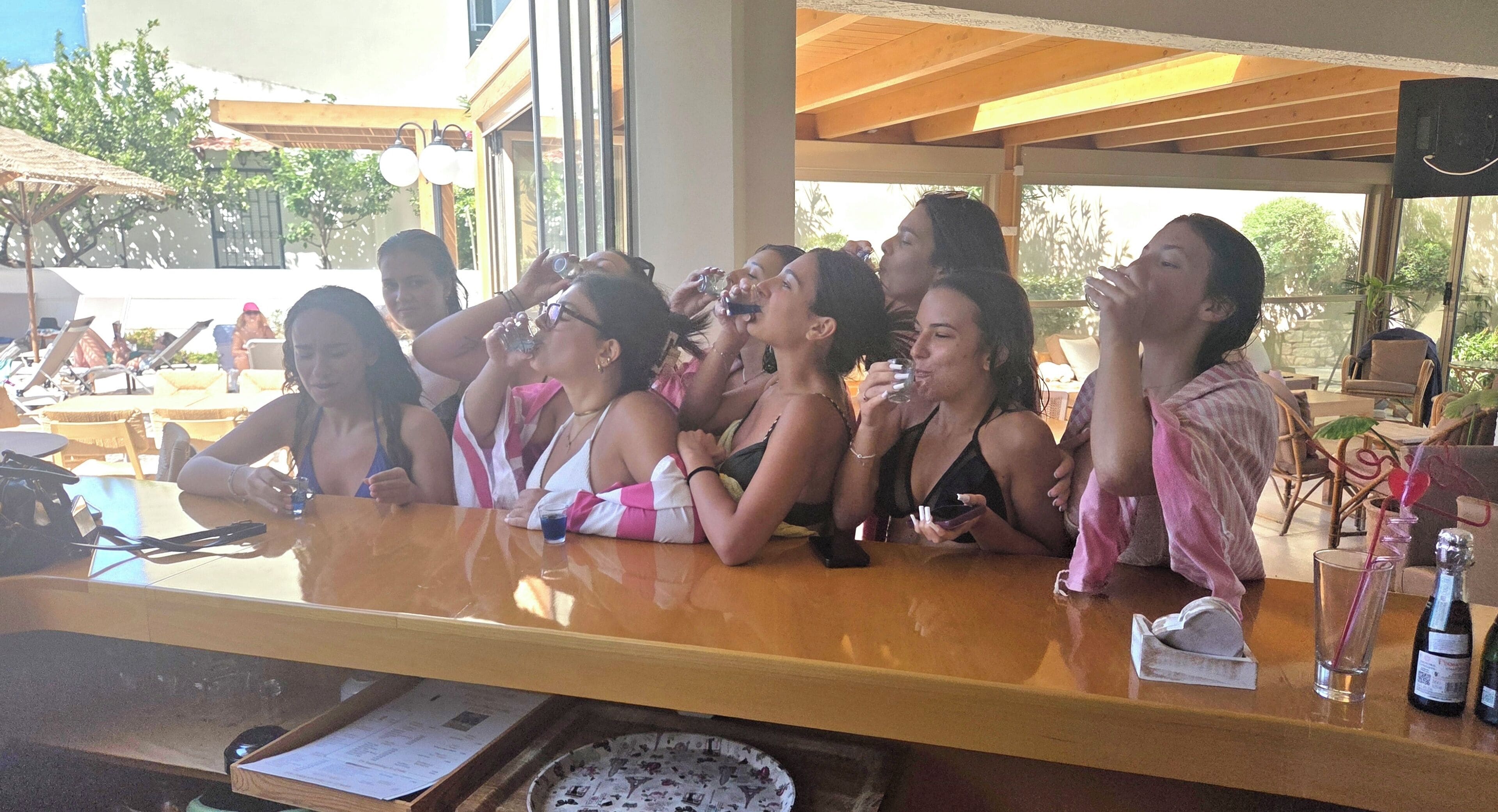 Un grupo de personas sentadas en un bar con mostrador de madera, mesas al aire libre y vistas al exterior.