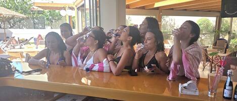 Un grupo de personas sentadas en un bar con mostrador de madera, mesas al aire libre y vistas al exterior.