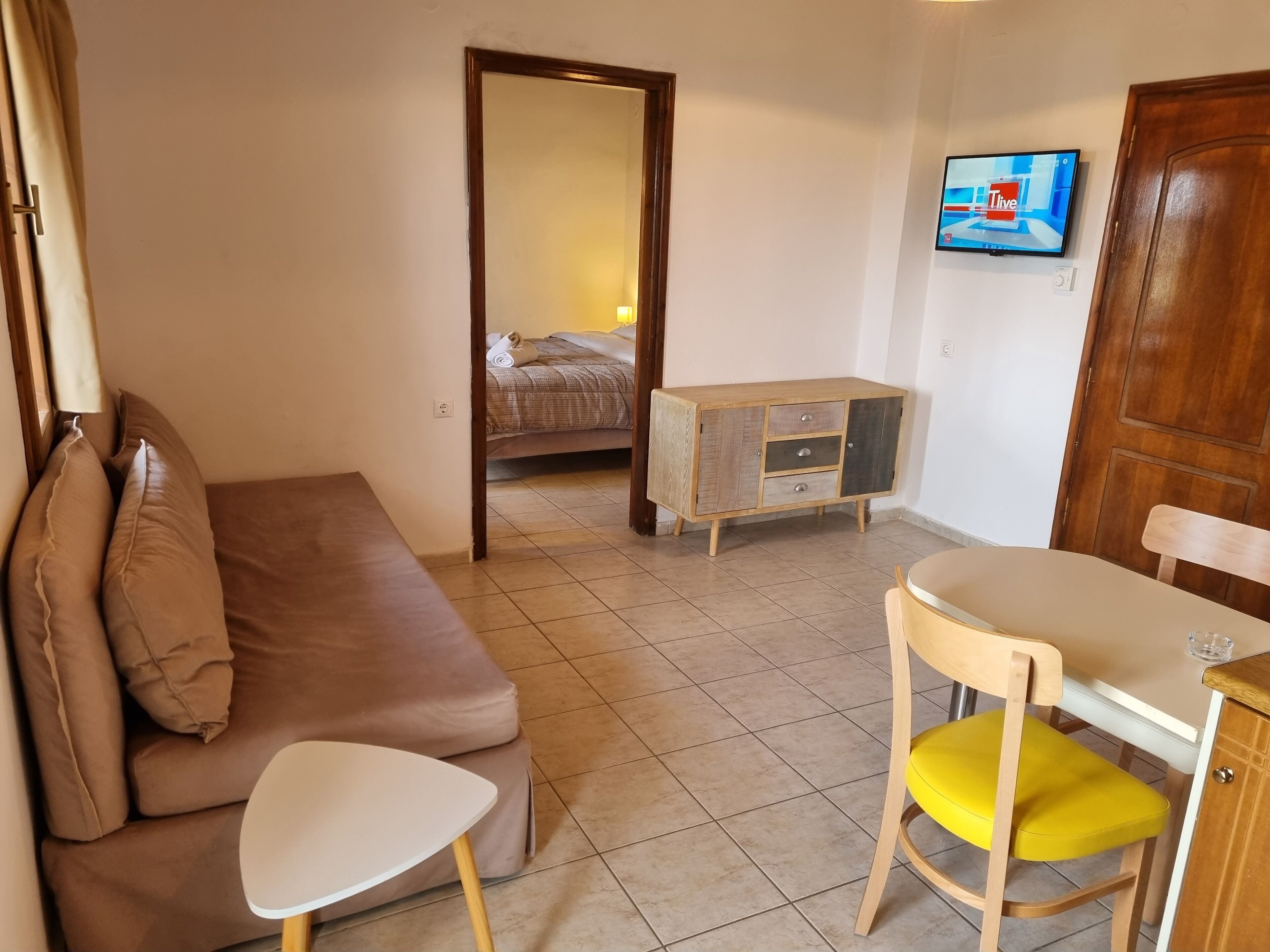 A compact room with a brown sofa, a small white table, a yellow chair, a wooden cabinet, and a bed in the background.