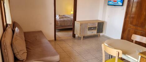 A compact room with a brown sofa, a small white table, a yellow chair, a wooden cabinet, and a bed in the background.